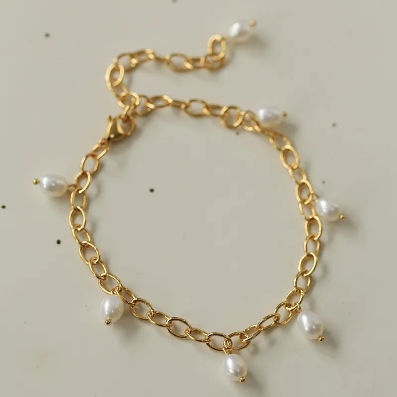 Bracelet de cheville doré à l'or avec une chaîne à maillons épais et plusieurs petites perles blanches nacrées pendantes. Le fermoir est de type mousqueton.