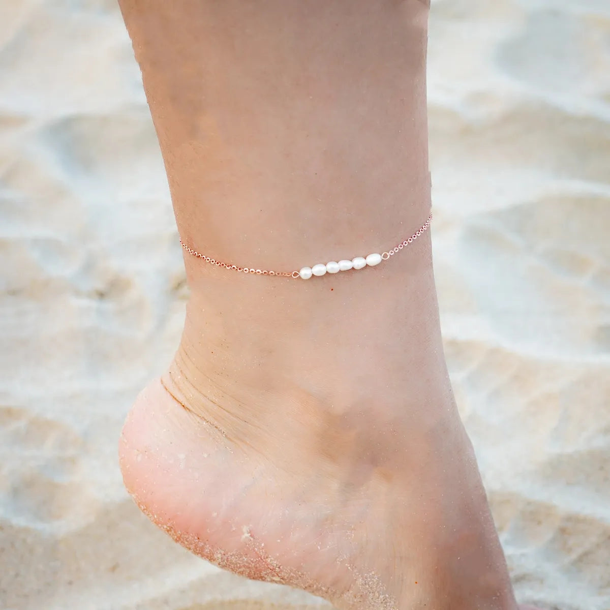 Bracelet de cheville délicat en argent avec une rangée de petites perles blanches sur une cheville posée sur du sable.