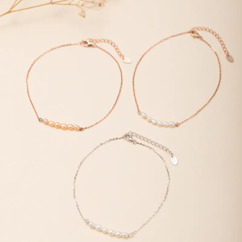 Trois bracelets de cheville en argent avec des perles blanches et roses disposées en ligne, sur fond clair.
