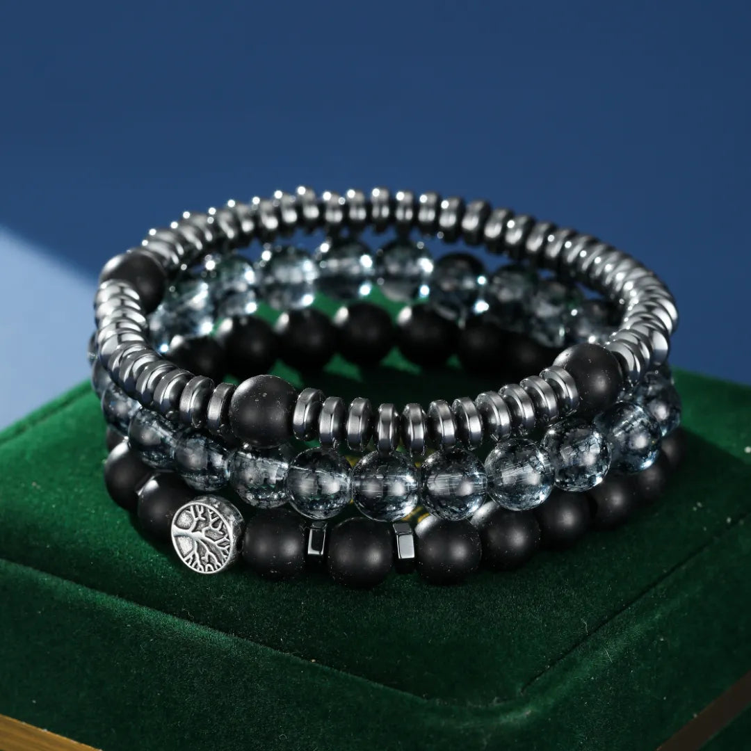Ensemble de trois bracelets pour hommes : un avec des perles d'hématite, un avec des perles d'obsidienne mate et des accents carrés, et un avec des perles transparentes et des accents métalliques. L'un des bracelets arbore un pendentif arbre de vie argenté.