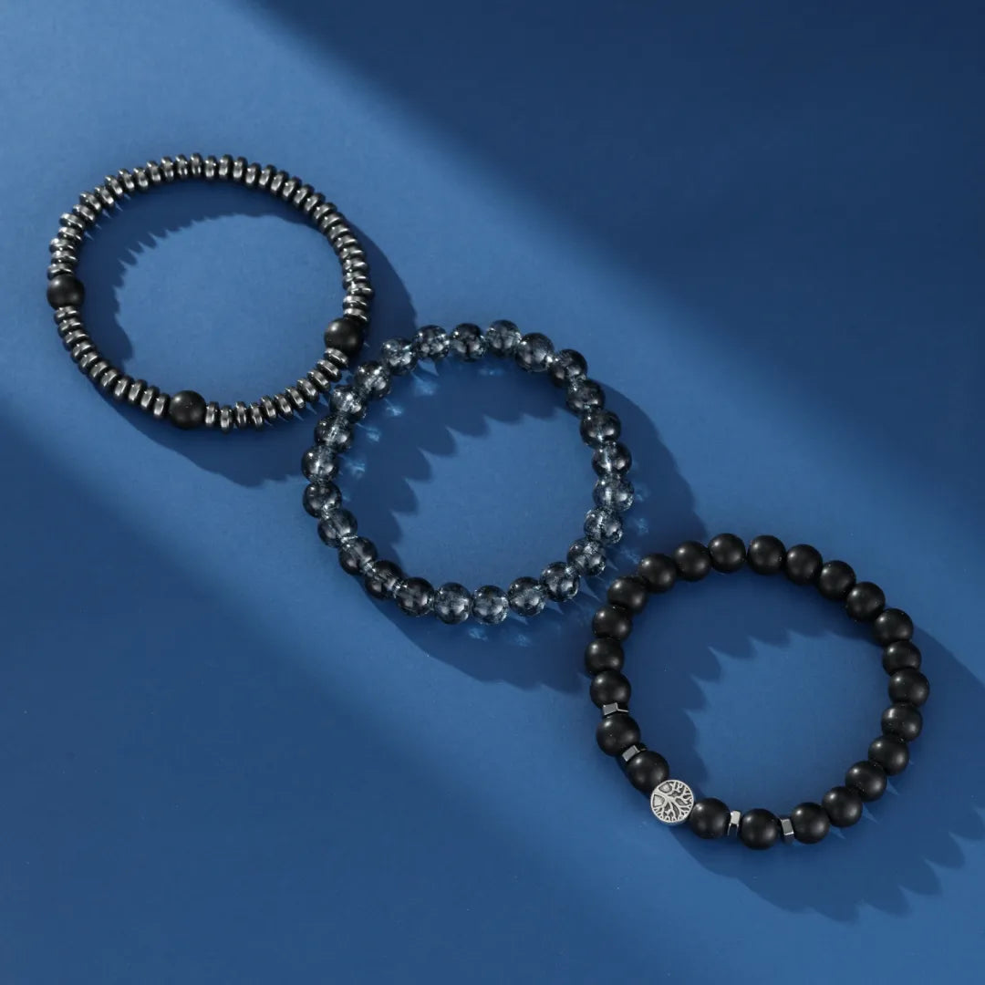 Trois bracelets pour hommes en perles sur fond bleu : un bracelet avec des perles d'hématite et d'obsidienne, un bracelet avec des perles grises facettées, et un bracelet en perles d'obsidienne noire avec un fermoir argenté orné d'un arbre de vie.