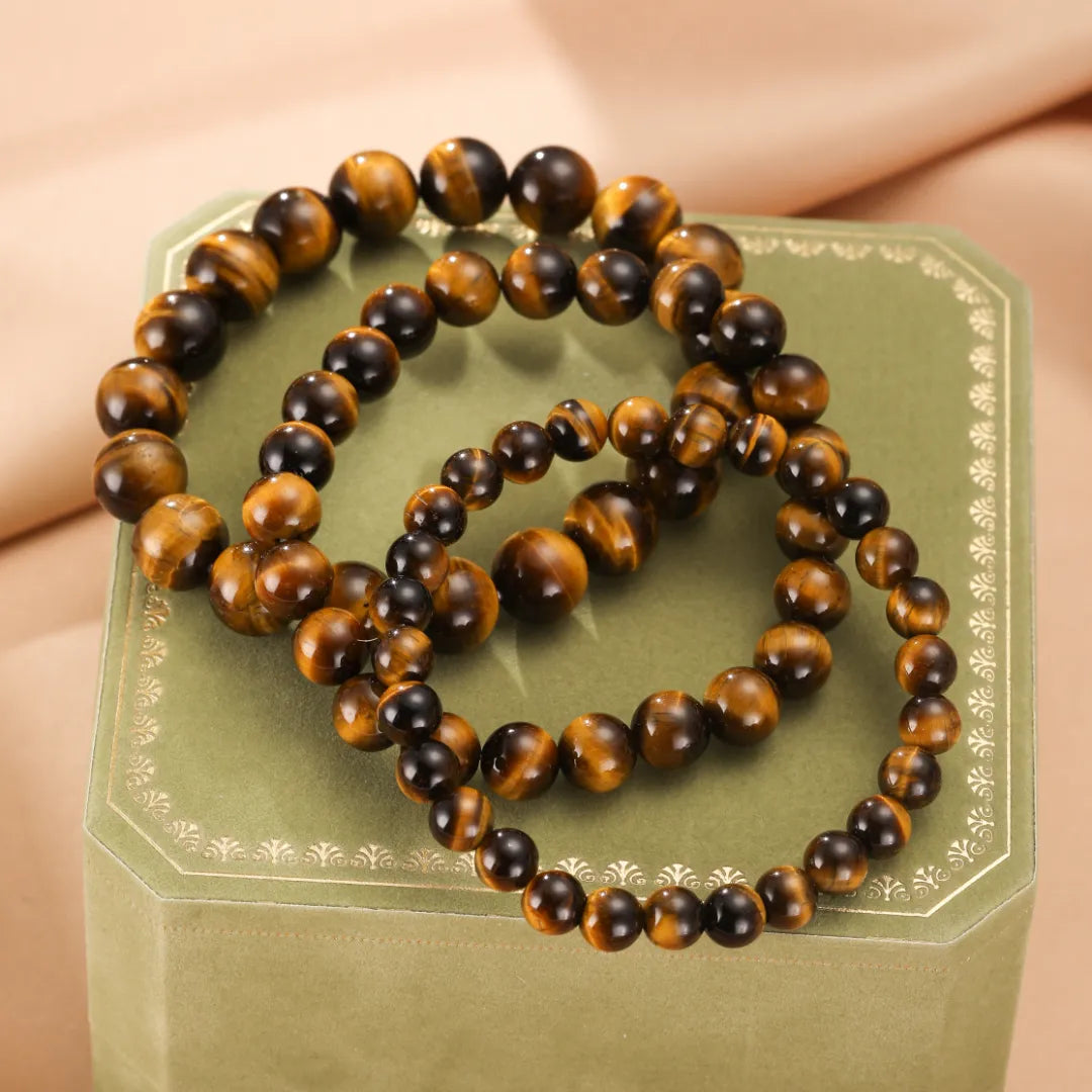 Trois bracelets d'œil de tigre "Force Pure" en perles rondes de tailles variées, posés sur une boîte verte décorée.