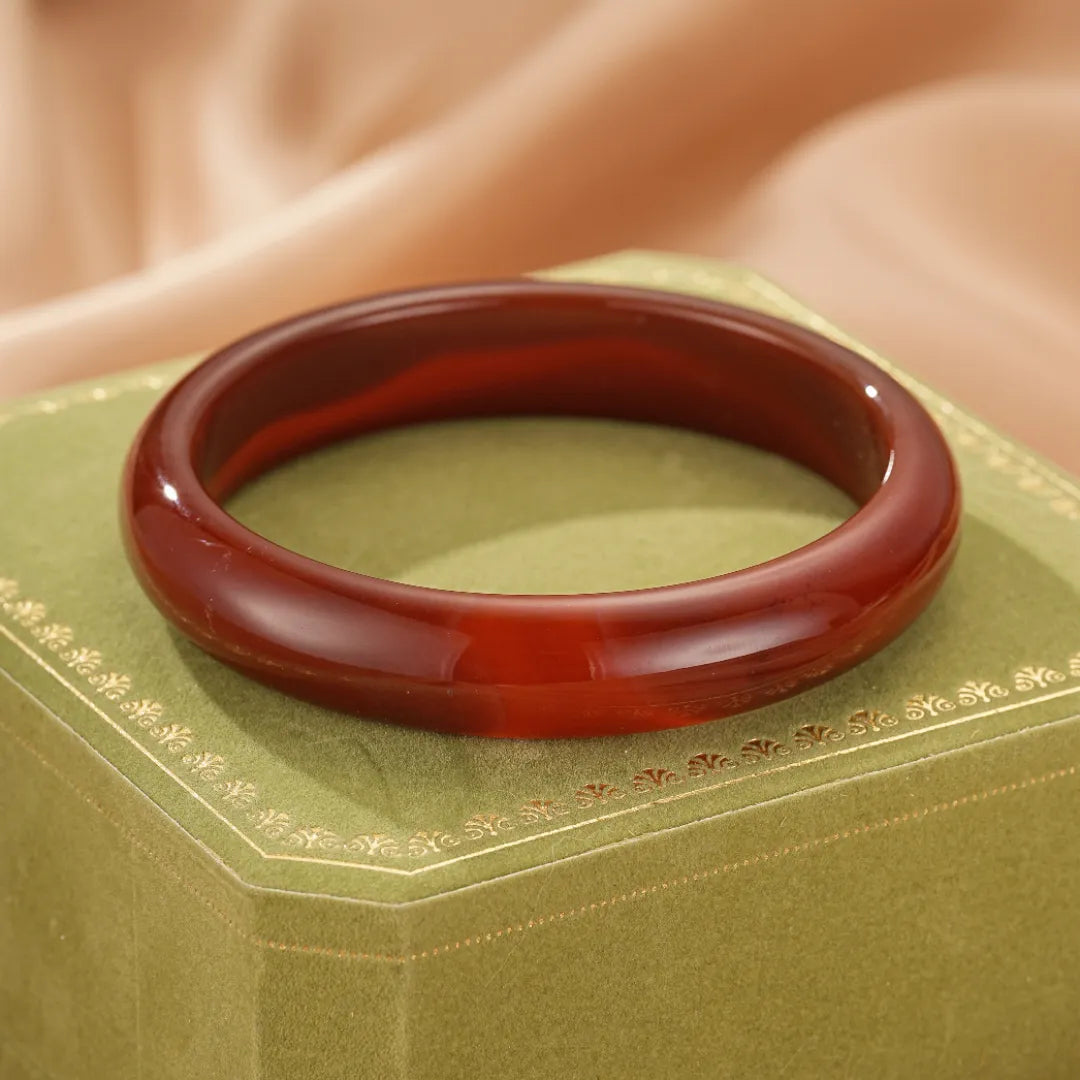 Bracelet jonc en agate rouge profond, poli et lisse, posé sur une boîte verte ornée de motifs dorés. Le bracelet dégage une impression de force et d'audace.