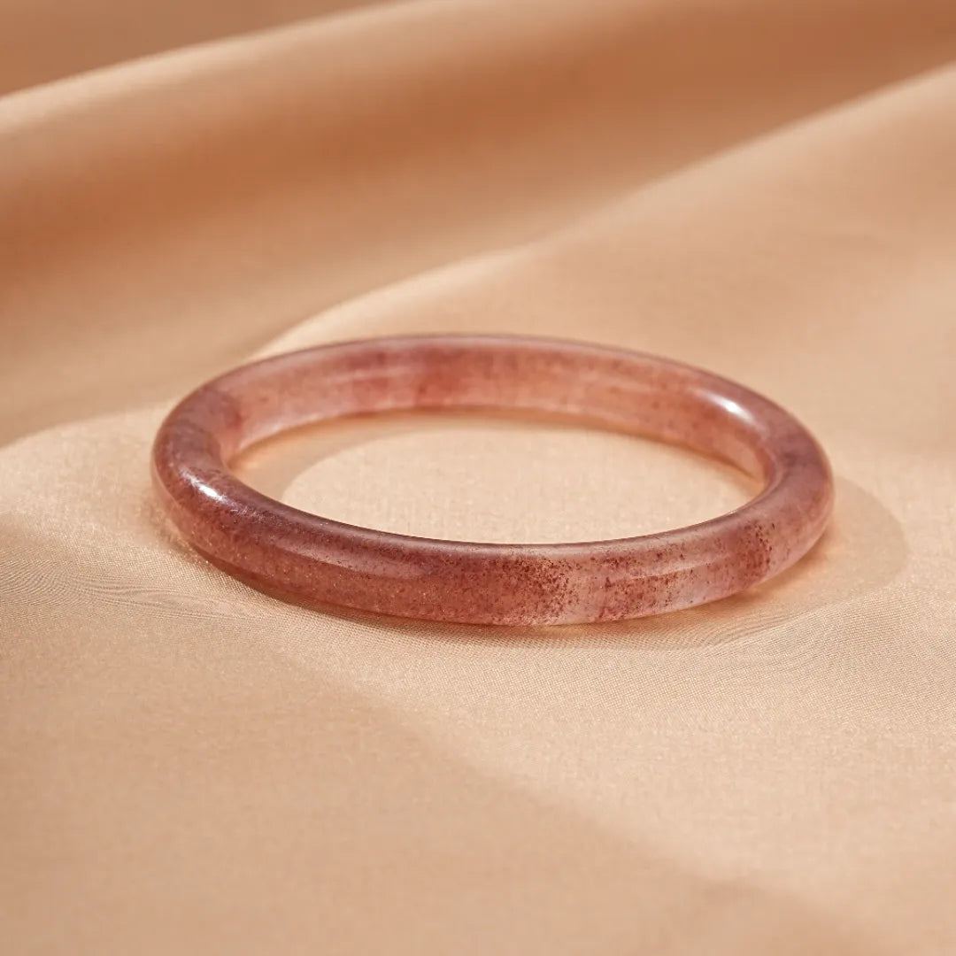 Bracelet jonc en quartz fraise rose translucide, nommé "Amour Joyeux", posé sur un tissu satiné de couleur pêche.