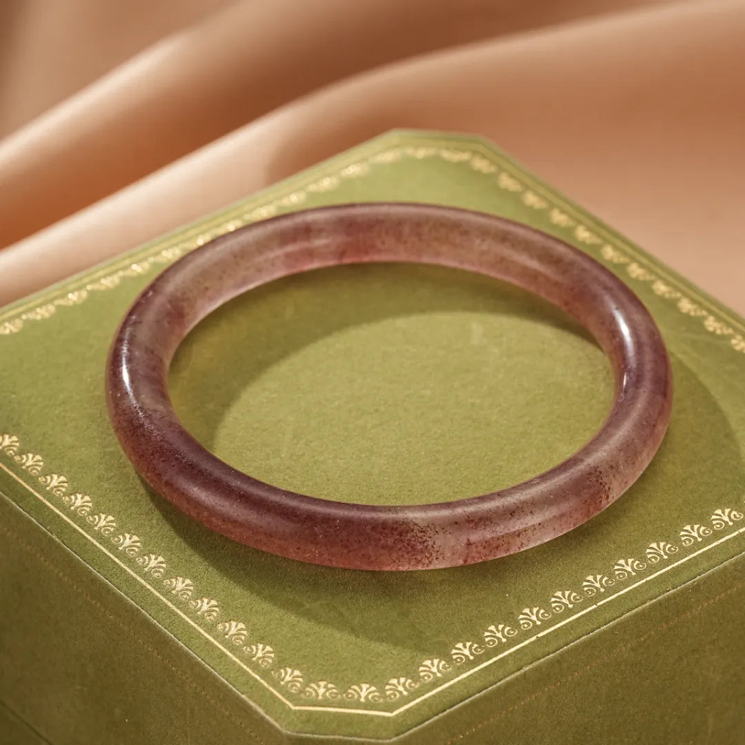 Bracelet jonc en quartz fraise "Amour Joyeux" de couleur violette, posé sur une boîte verte ornée de motifs dorés.