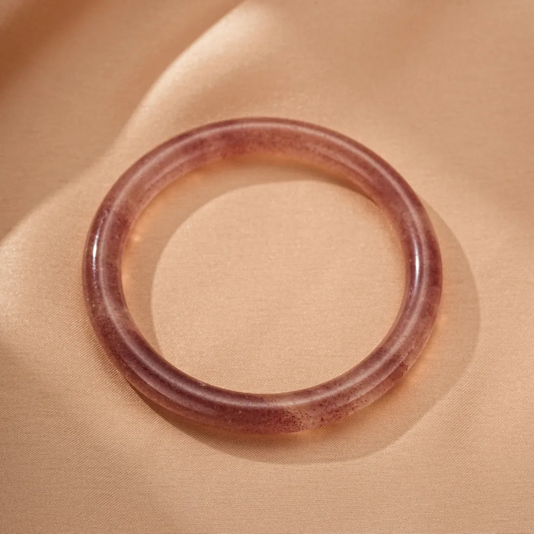 Bracelet jonc en quartz fraise transparent et rose, style "Amour Joyeux", posé sur un fond satiné beige.