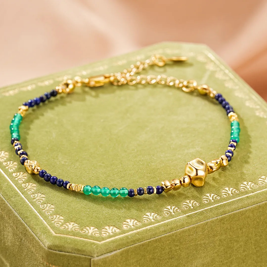 Bracelet en argent doré avec perles de lapis-lazuli bleues, de jade vert et un cube doré au centre, le bracelet "Sagesse Intérieure" est posé sur une boîte verte.