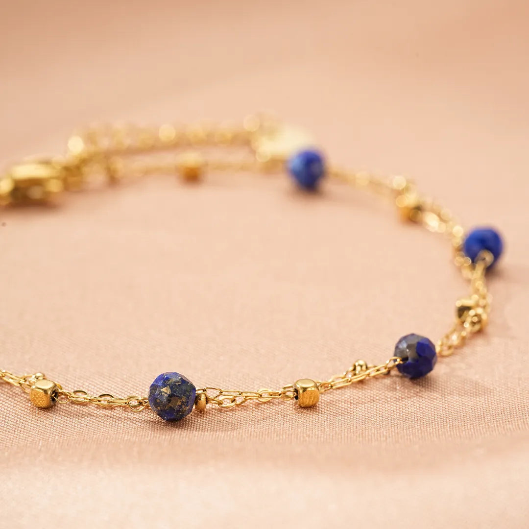 Bracelet fin en plaqué or orné de perles d'amazonite ou de lapis-lazuli et de petits cubes dorés, sur fond rose poudré.