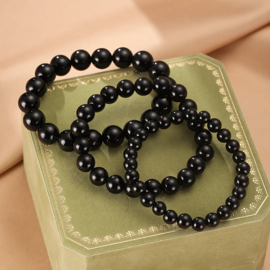 Trois bracelets en perles d'obsidienne noire disposés sur une boîte verte ornée de motifs dorés.
