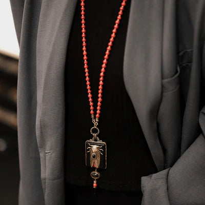 Collier fantaisie avec pendentif cigale dorée et agate sur chaîne de perles rouges corail, porté sur un pull noir sous une veste grise.