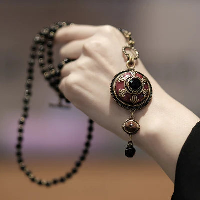 Collier "Merveille d'Antan" avec un pendentif rond rouge et or orné d'une agate noire et de pierres marron, porté sur une chaîne de perles noires.