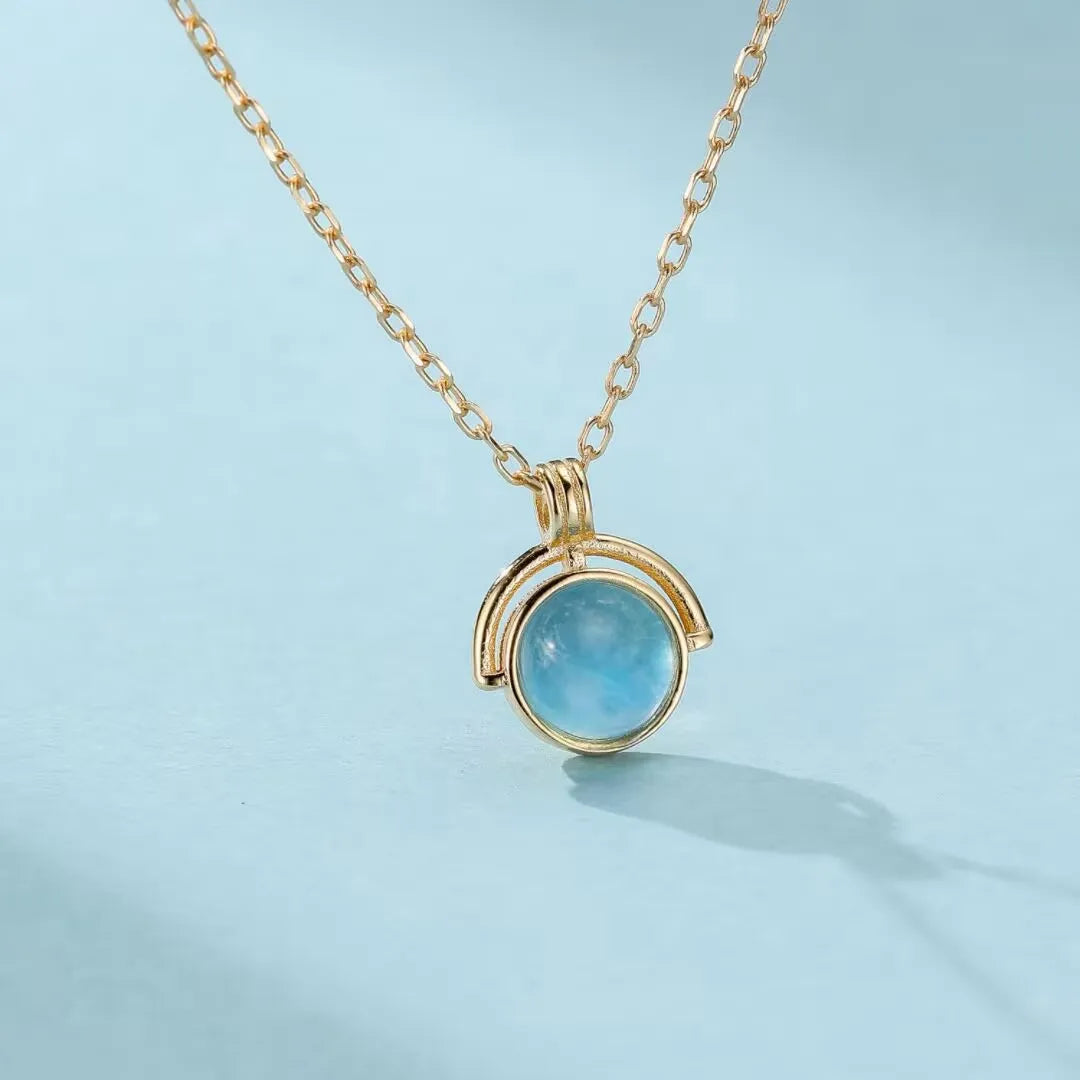 Collier plaqué or avec une pierre d'aigue-marine bleue ronde suspendue dans un cercle ouvert, sur fond bleu clair.