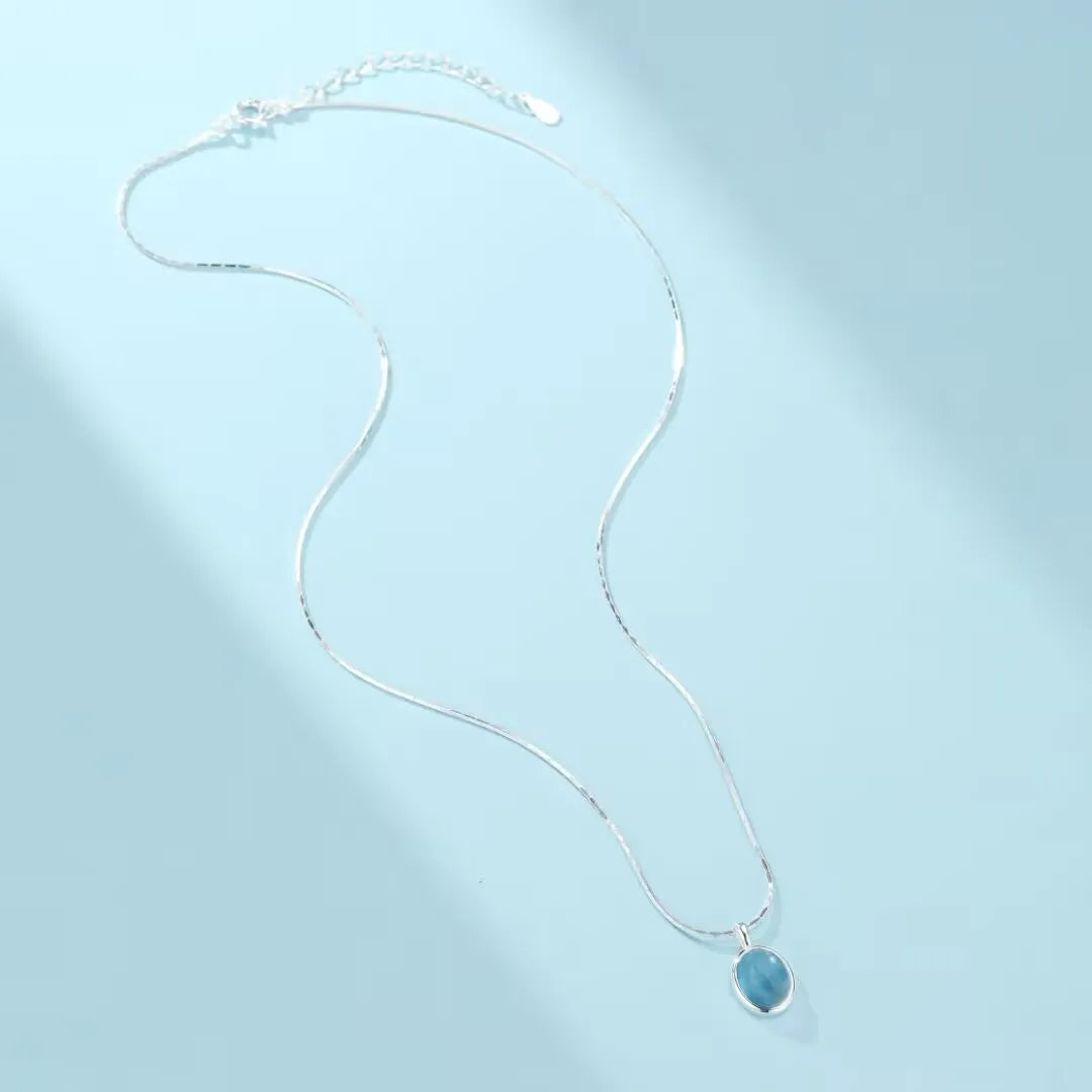 Collier en argent 925 avec un pendentif ovale en aigue-marine bleu clair, sur fond bleu pastel.