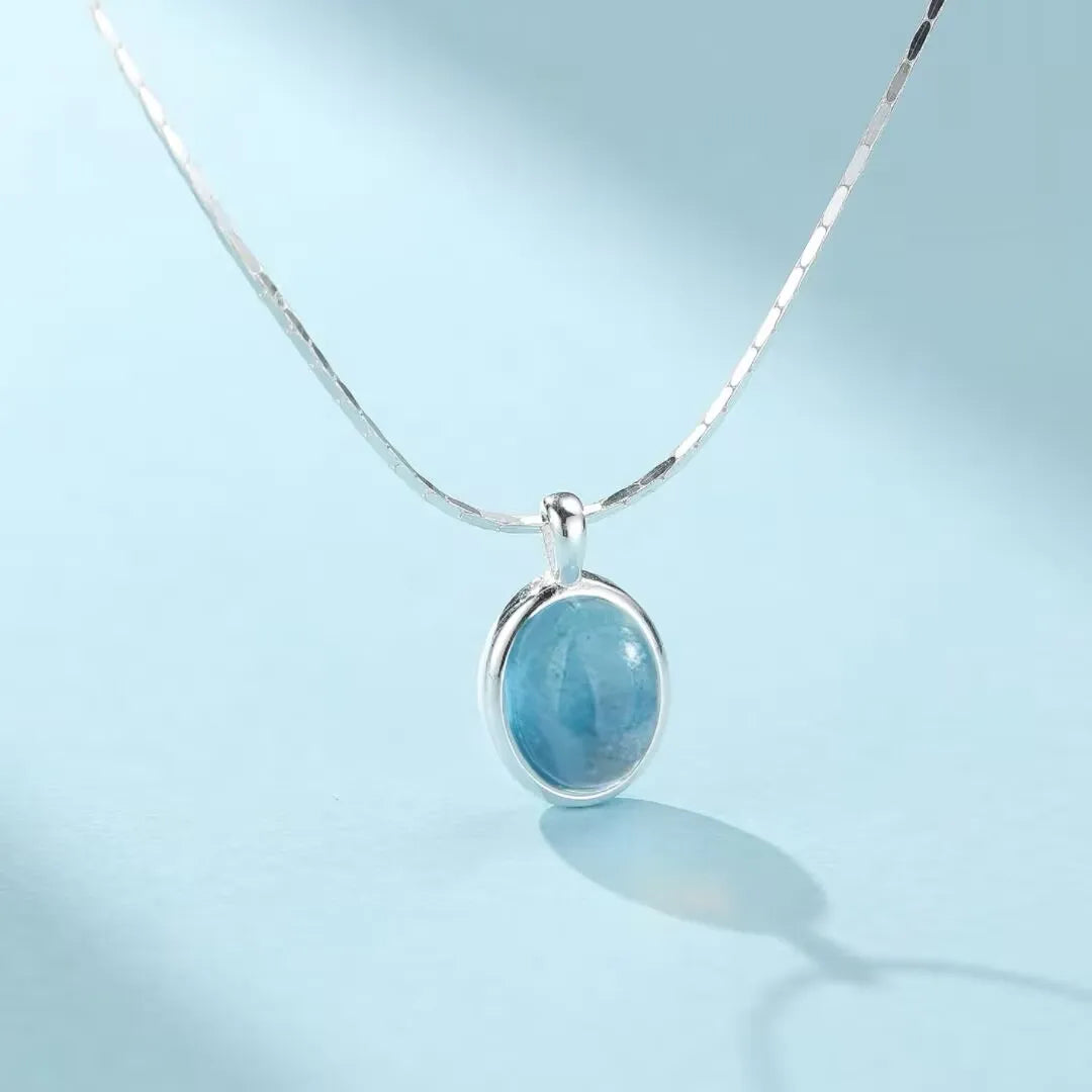 Collier en argent 925 avec un pendentif ovale en aigue-marine bleue, sur fond bleu clair.