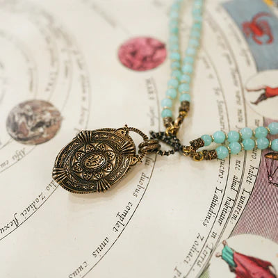 Collier fantaisie "Voyage Marin" avec un pendentif ovale ouvragé couleur bronze et des perles d'amazonite bleu clair, posé sur une carte ancienne.