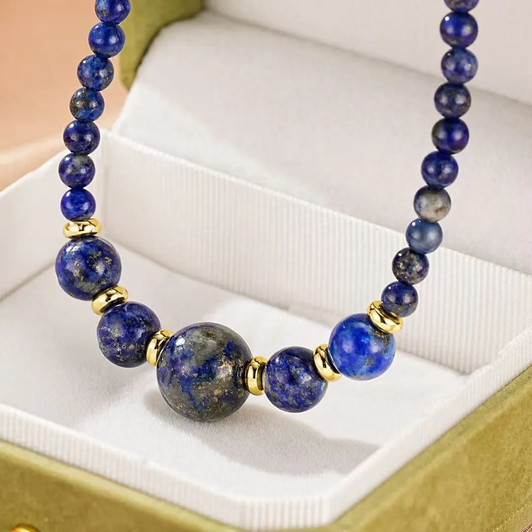 Gros plan sur un collier en pierres de lapis-lazuli bleues avec des perles de tailles différentes et des accents dorés, présenté dans une boîte cadeau verte.