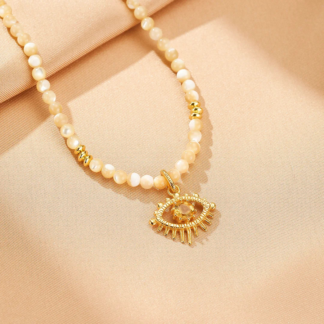 Gros plan sur un collier ras de cou fait de perles en nacre de couleur crème et d'une breloque œil stylisée en argent doré, ornée d'une petite pierre claire au centre.