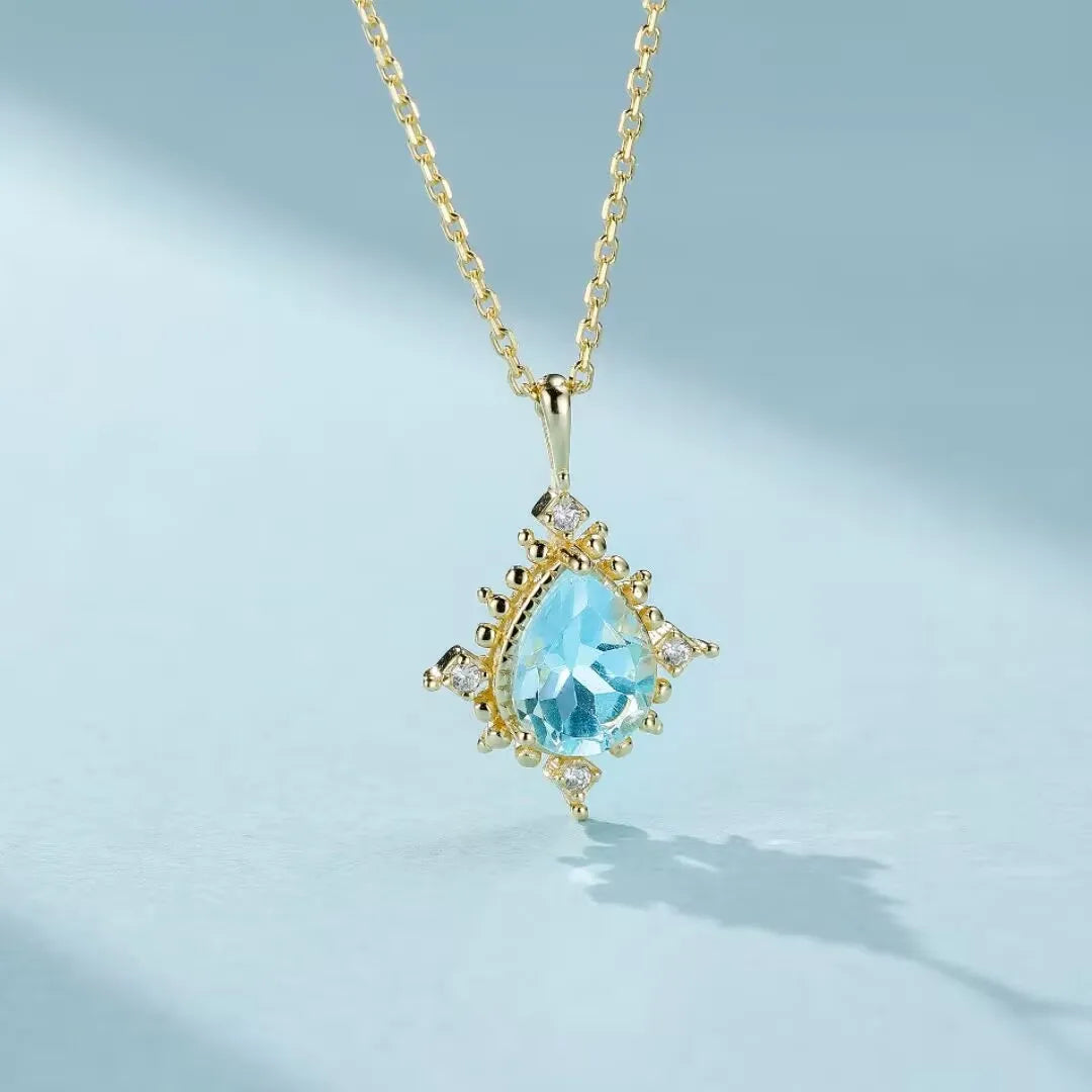 Collier avec pendentif en forme de goutte d'eau en topaze bleue serti sur un cadre en argent 925 doré, orné de petites pierres scintillantes.