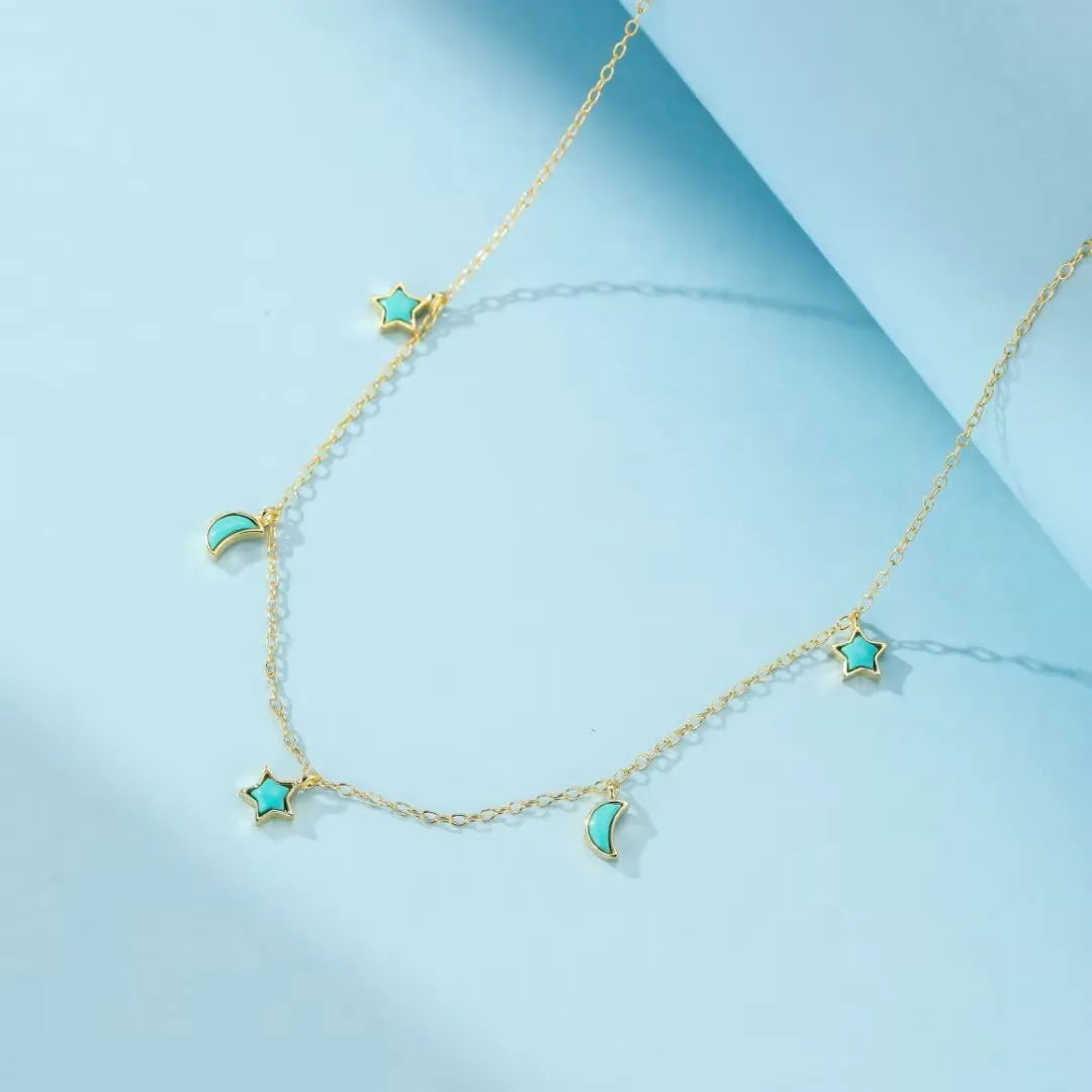 Collier "Équilibre Céleste" en argent doré à l'or, orné de petites étoiles et lunes en turquoise bleu clair, sur fond bleu pâle.