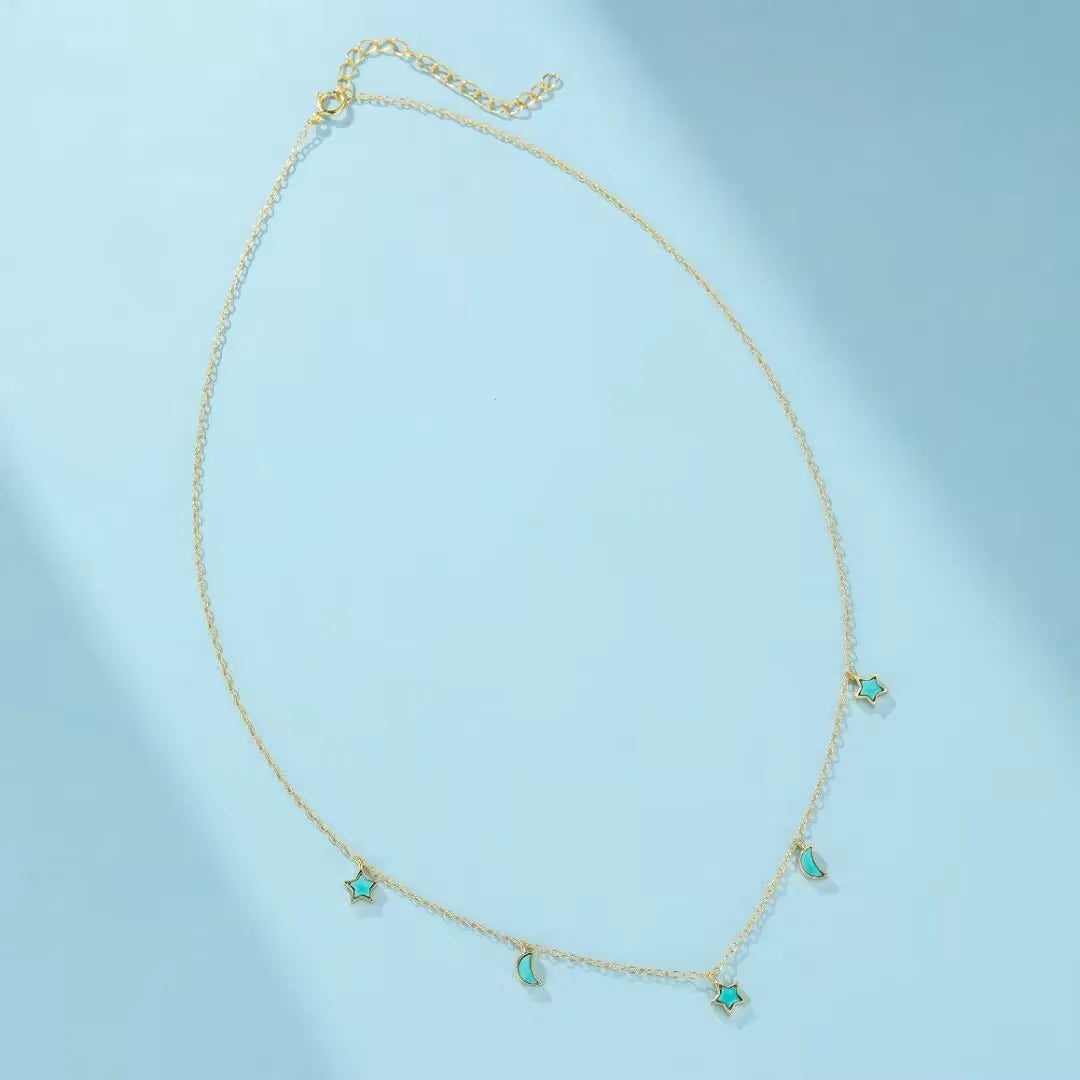 Collier "Équilibre Céleste" en argent doré, orné de petites étoiles et lunes en turquoise, sur fond bleu clair.
