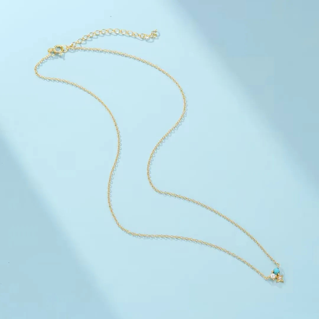 Collier fin en argent doré or, orné d'une petite turquoise et d'une perle nacrée, le bijou "Sagesse Stellaire" est présenté sur un fond bleu clair.