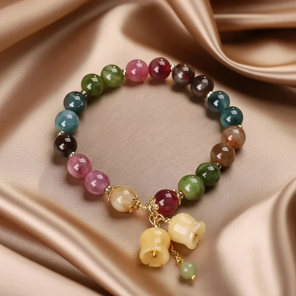 Bracelet "Harmonie" en pierres naturelles multicolores des 7 chakras, orné de breloques en forme de clochettes et d'une petite pierre verte, sur fond satiné.
