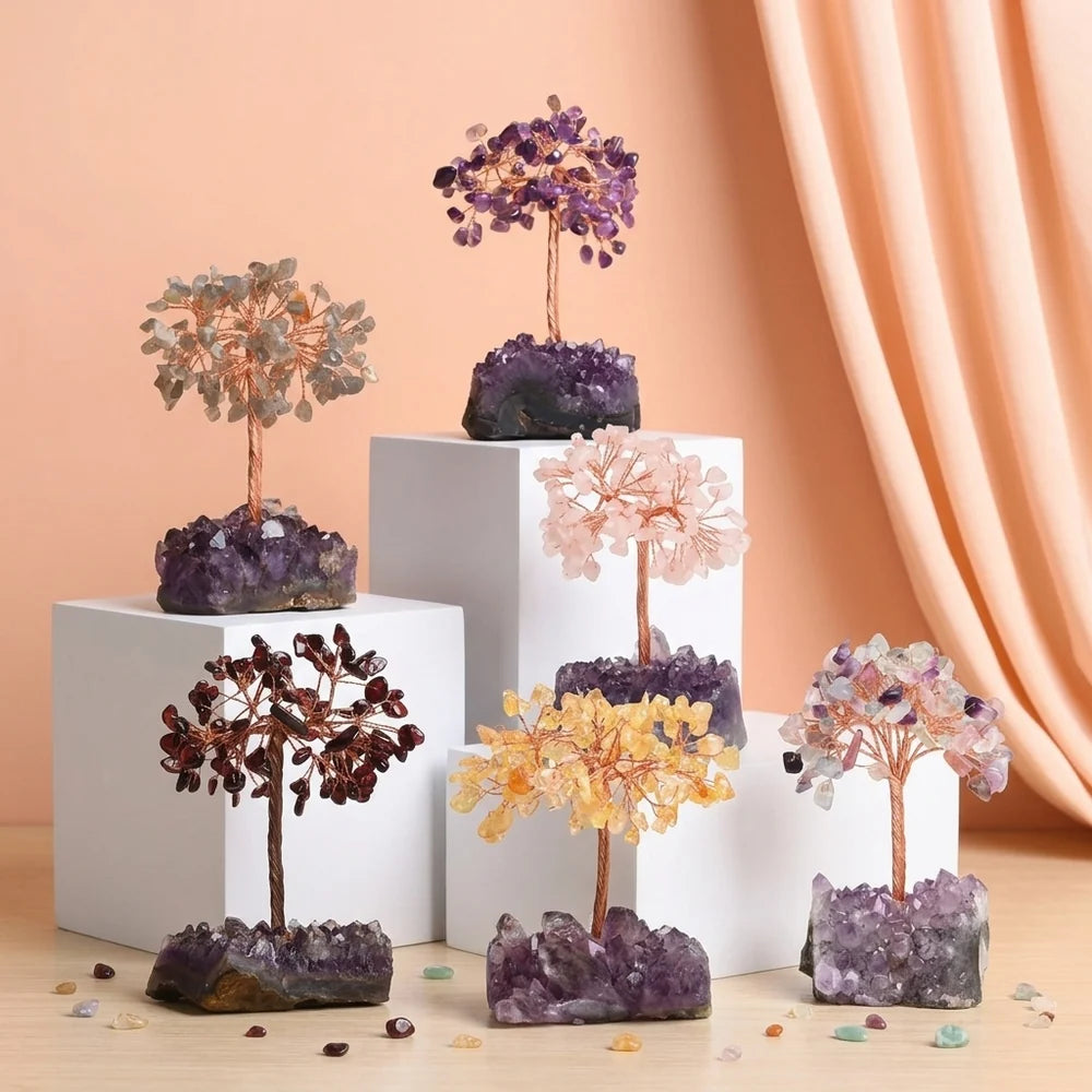Cinq arbres de vie décoratifs en pierres naturelles de différentes couleurs (améthyste, quartz rose, citrine, grenat, et mélange) sur des bases d'améthyste brutes, disposés sur des cubes blancs et une table en bois.